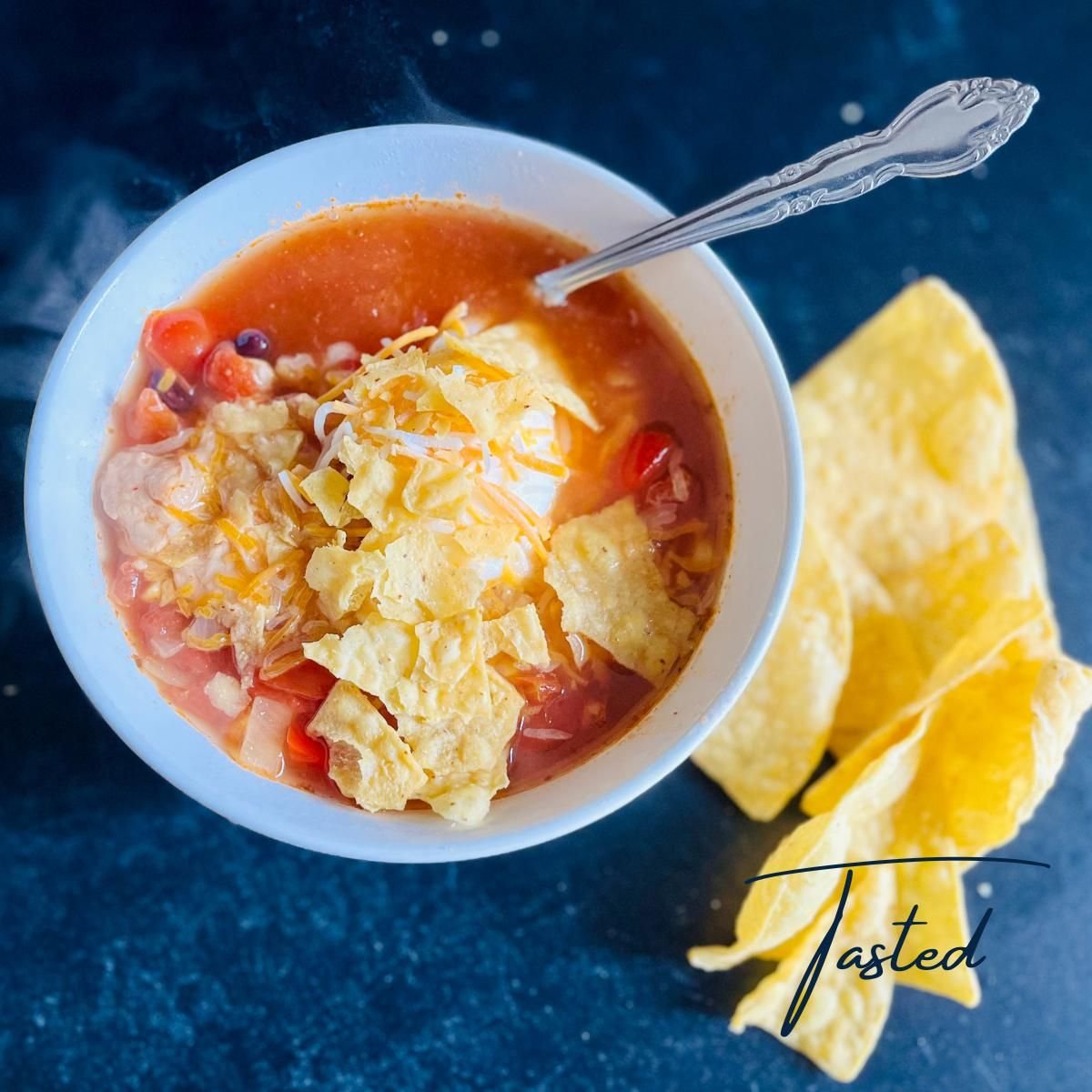 Bowl of chicken taco soup topped with crushed tortilla chips, shredded cheddar and sour cream, with tortilla chips on the side
