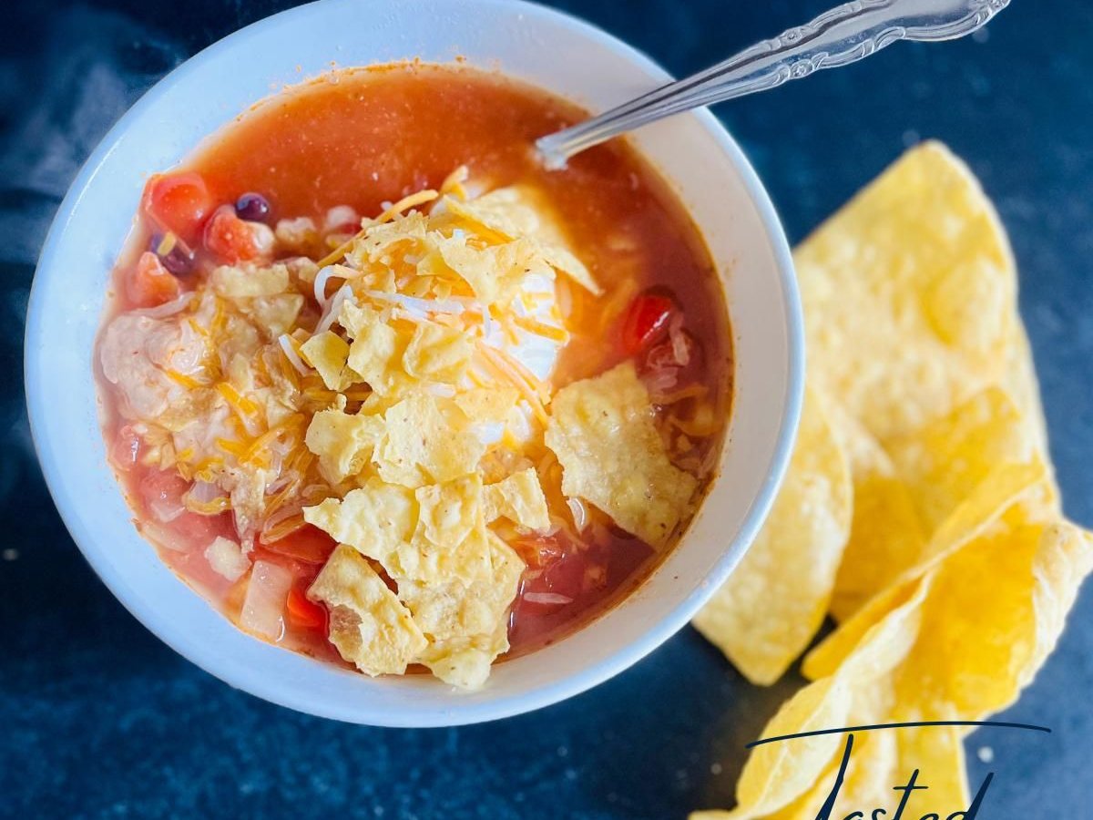 Bowl of chicken taco soup topped with crushed tortilla chips, shredded cheddar and sour cream, with tortilla chips on the side