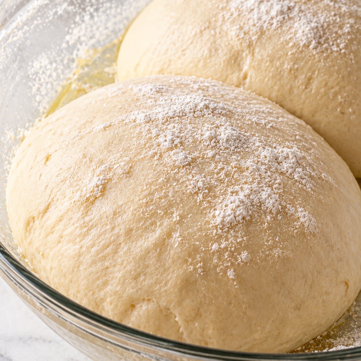Two risen balls of pizza dough dusted with flour in a glass bowl
