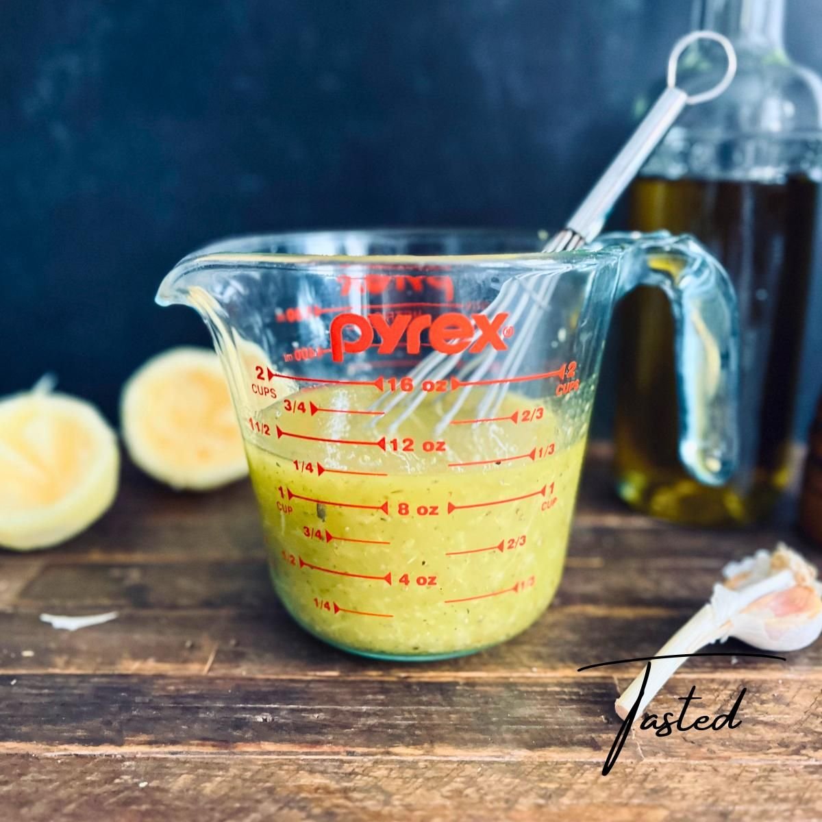 Greek lemon dressing in a glass measuring cup with a whisk, next to lemon halves, garlic and a bottle of olive oil