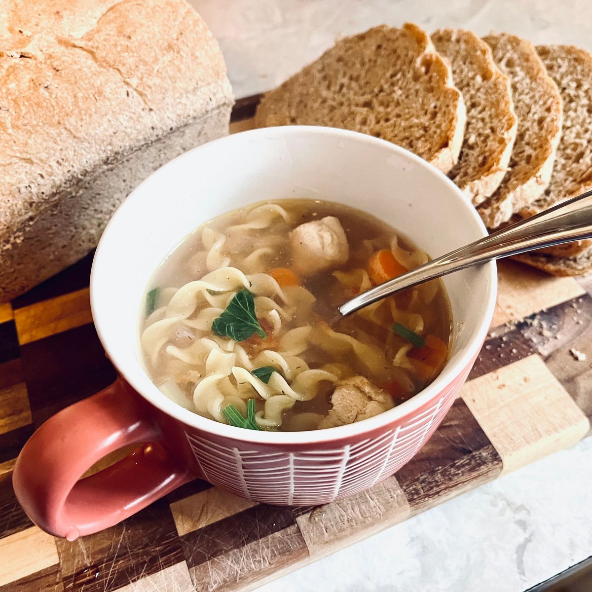 Mug of chicken noodle soup with carrots and herbs on a wooden board, with a loaf of homemade bread and slices behind it