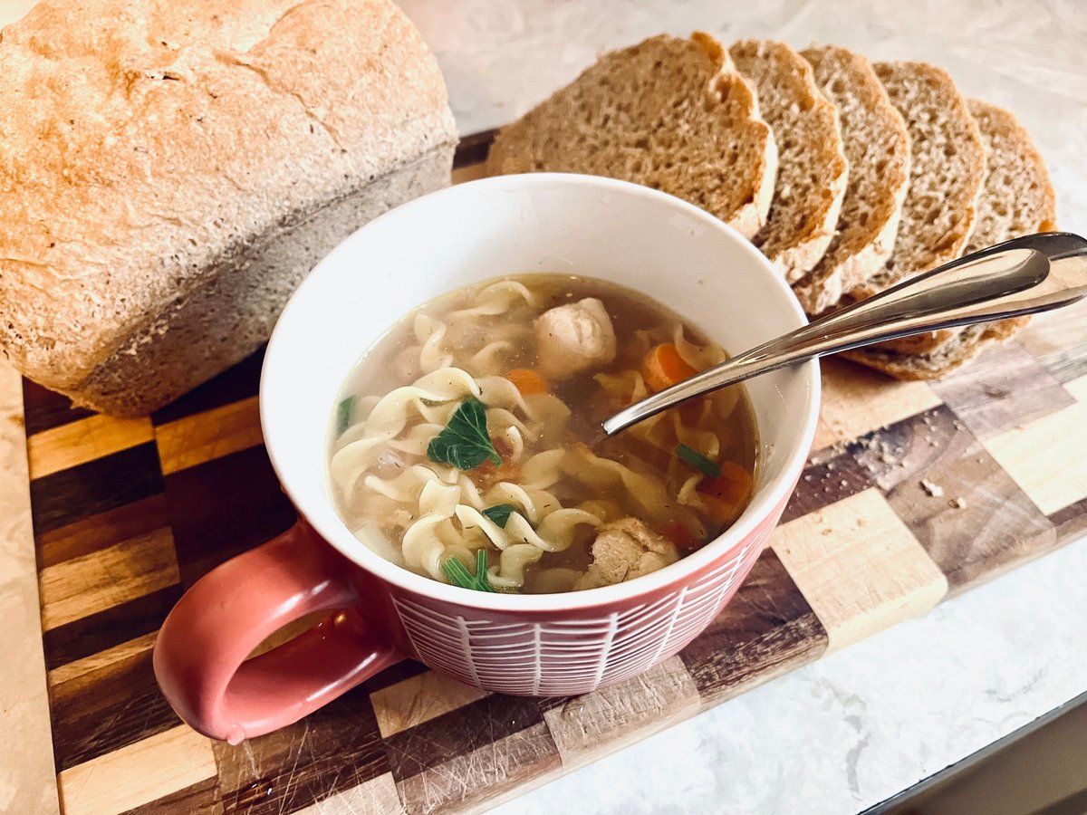 Mug of chicken noodle soup with carrots and herbs on a wooden board, with a loaf of homemade bread and slices behind it
