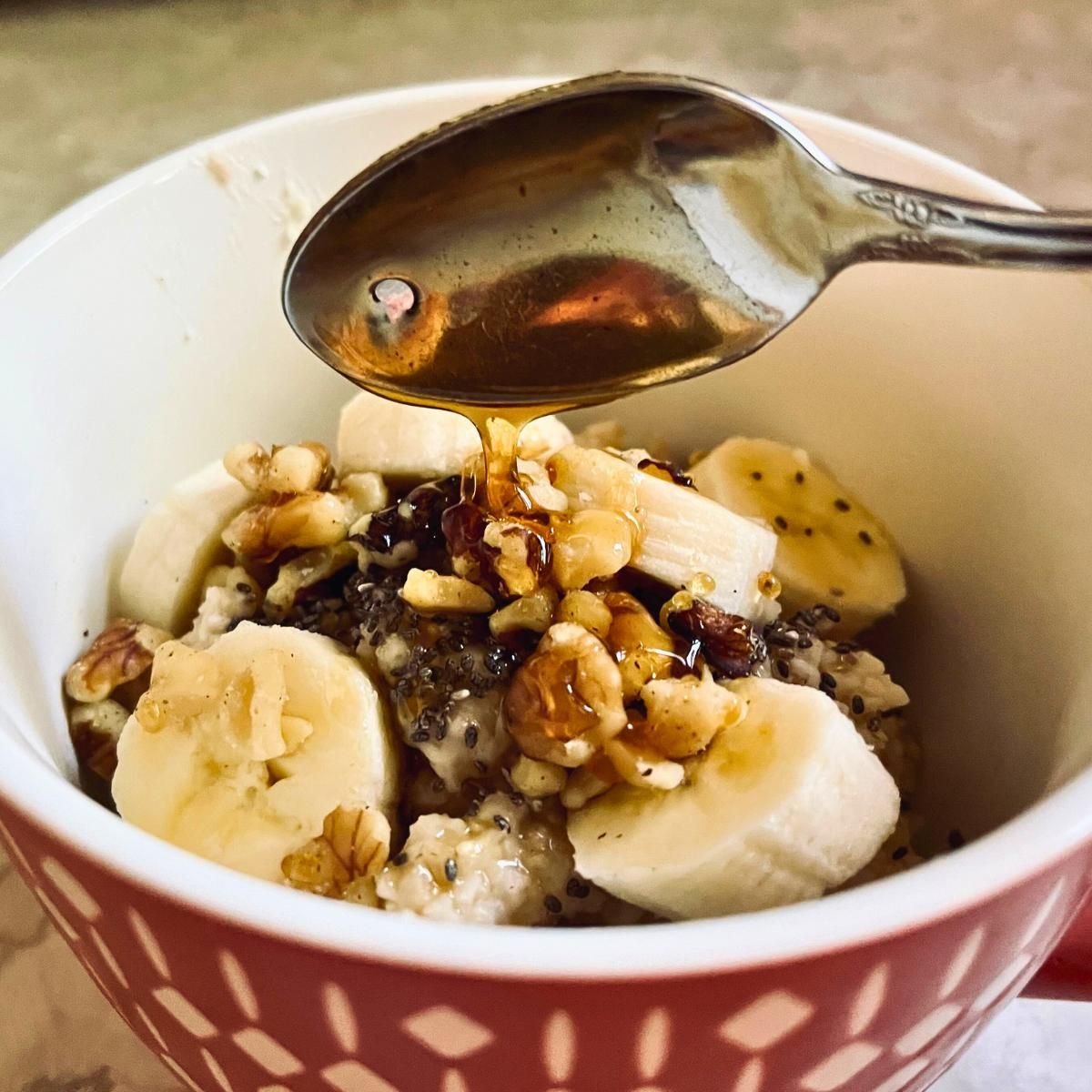 Bowl of soaked oatmeal porridge topped with banana slices and walnuts, with honey drizzled from a spoon