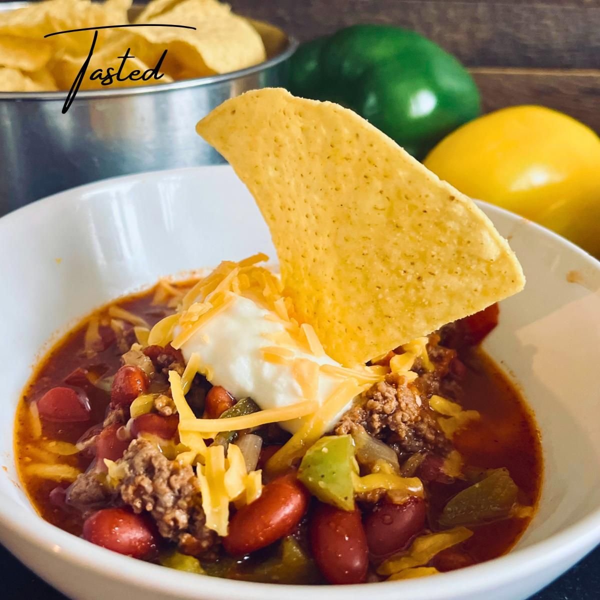 Bowl of beef chili with kidney beans and peppers, topped with sour cream, shredded cheddar and a tortilla chip