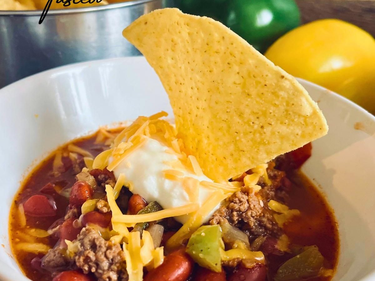 Bowl of beef chili with kidney beans and peppers, topped with sour cream, shredded cheddar and a tortilla chip