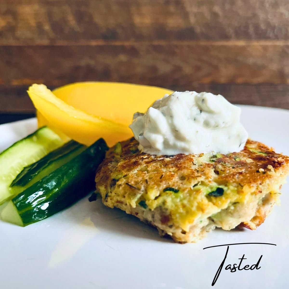 Zucchini tuna patty topped with a dollop of herbed sauce, with cucumber spears and mango slices